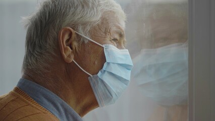 Old man with mask face in home looking out window, portrait reflected on glass