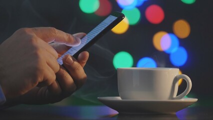 Detail of hand with smartphone near steaming coffee cup