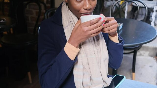 Happy African American woman sitting in cafe drinking coffee smiling thinking looking around tourist autumn coffee break cloae up