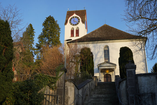 Protestant church in Hinwil, Switzerland captured in front low angle view with its clock tower. There is a staircase leading to the entrance. Trees are around it and clear blue sky on the background.