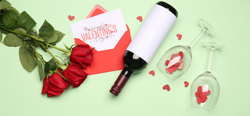 Fototapeta na wymiar Bottle of wine, greeting card in envelope, rose flowers and glasses on green background. Valentine's Day celebration