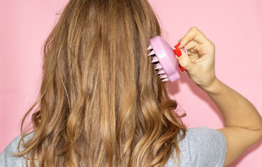 blonde hair girl woman holding ponytail up or silicone soft shampoo brush with bristles isolated on pink.hair growth stimulating scalp massage healthy long curly hair.young woman back view hands up