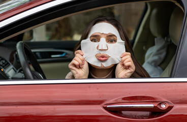 woman with facial sheet textile mask on steering wheel in driver seat looking in rear view mirror making crazy funny faces kissing gesture.skin face care in hurry concept red color auto