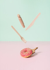 Sweet pink donut ready for taste test on kitchen board. Flying fork and knife above dessert. Creative eating concept on pastel green background. Minimal treat concept.