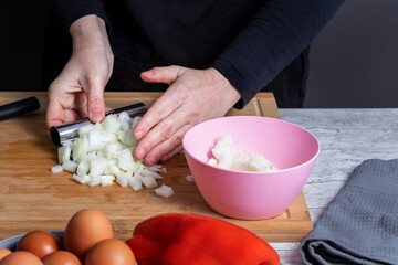 Hands of working woman picking up cut onion on a kitchen board to put it in pink boll. In front of defocused red pepper and gray boll with eggs. Cooking concept.