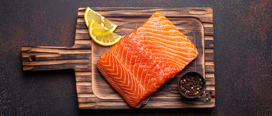 Fresh raw salmon fillet on wooden kitchen cutting board with seasonings and lemon top view on dark...