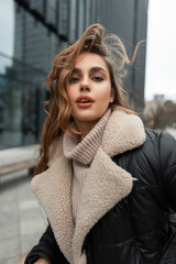 Fototapeta na wymiar Beautiful fashionable woman model with make-up and curly hairstyle in stylish winter wear with knitted beige vintage sweater and fashion trendy jacket walks in the modern city