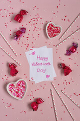 Vertical Valentine's Day background. Congratulations on Valentine's Day written on a white sheet of paper surrounded by ceramic hearts with sprinkles and chocolates on pink.