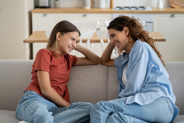 Cheerful friendly family of woman and teenager daughter are sits on sofa and laughing discussing funny school stories. Positive smiling mom and little girl spend time together gossip on weekend  - obrazy, fototapety, plakaty