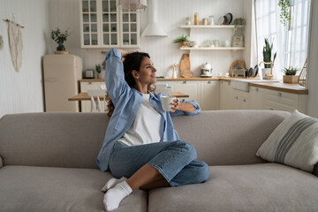 Happy dreamy young woman wearing casual clothes resting with cup of tea at cozy home, smiling...