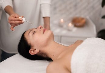 Obraz na płótnie Canvas Young lady lying closed eyes while cosmetician applying essential oil on face