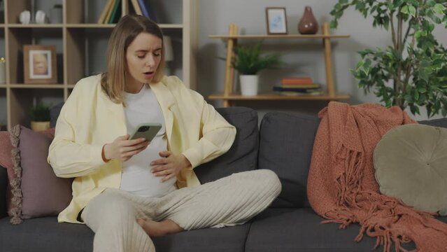 Pregnant caucasian woman feeling labor contractions while searching information about babies on smartphone. Concept of maternity and technology.