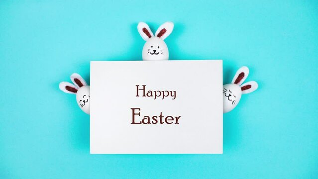 4k Three Easter bunnies made of white chicken eggs emerge from behind a sheet of white paper. Holiday greeting card "Happy Easter". Blue background. Bunny Day. Stop motion animation. Copy space.