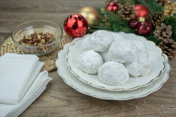 Snowball cookies presented on beautiful white plates surrounded by christmas ornaments, white napkin and small bowl of nuts. Pine cones.  Wooden surface. Powdered sugar.