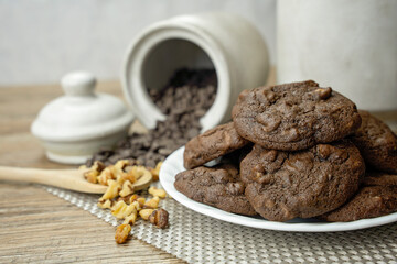 Double chocolate cookies freshly baked on a white dish with chocolate chips, walnuts, pecans and wooden spoon.  