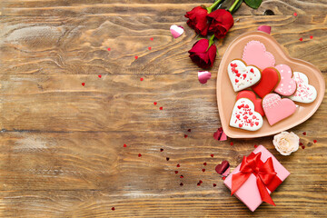 Composition with plate of tasty heart shaped cookies, gift box and rose flowers on wooden background. Valentine's Day celebration