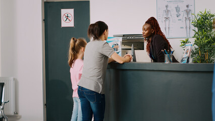 African american receptionist giving assistance to mother and kid, talking about doctor appointment at hospital reception counter. Medical staff filling in checkup report papers in facility lobby. - obrazy, fototapety, plakaty