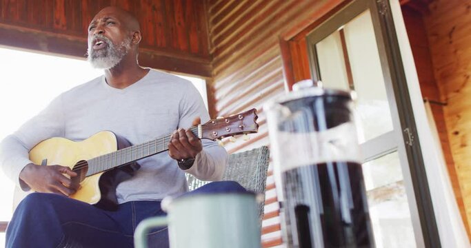 Happy senior african american man in log cabin, playing guitar on balcony, slow motion