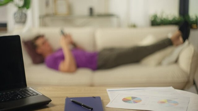 Businessman took a break in the home office and lies with the phone on the sofa. The man uses the Internet and texts. A hard day's work at home. Table with coffee, computer and documents.