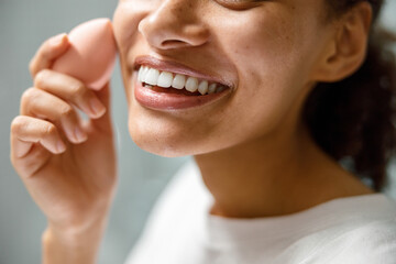 Close up of smiling african woman with sponge applying makeup at home bathroom