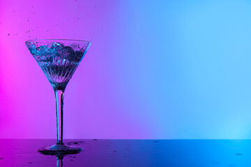 Glass of martini and splashes on table in neon lights, space for text