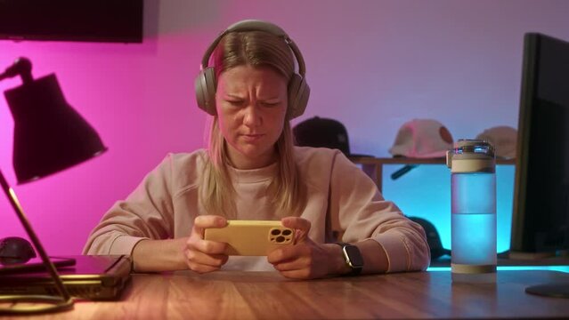 young woman sits at table at home in headphones and play game on phone. neon light in room illuminates place of streamer or avid gamer girl. gamer is passionate about playing on smartphone.