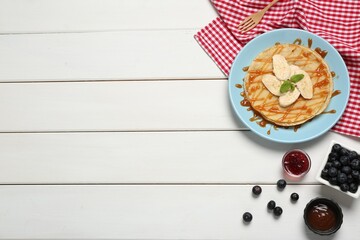 Delicious crepes with blueberries, mint, banana and jam on white wooden table, flat lay. Space for text