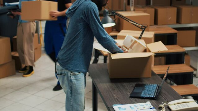 African american man putting products in cardboard boxes, preparing order for shipment. Young person doing quality control to ship merchandise to customers from storage room space.