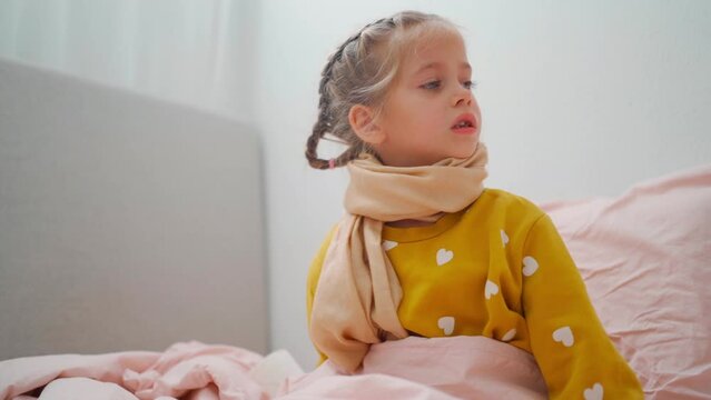 Child girl in plaid and scarf suffering from Coronavirus or allergy, sneezes snot into napkin on sofa at home 