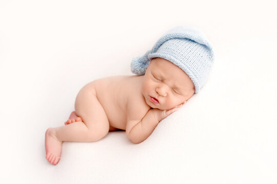 Cute newborn baby boy in the first days of life, sleeping naked on white fabrics in a knitted woolen blue cap. Studio professional macro photography, portrait of a newborn. Image with selective focus.