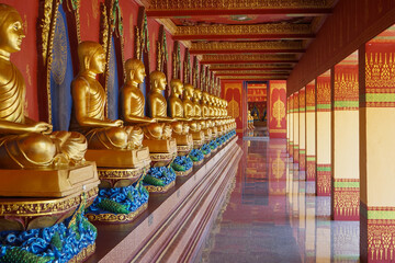 Selective focus on the golden Buddha statue sits among the defocused statues along the corridor at the Buddhism temple with defocused walking people in background
