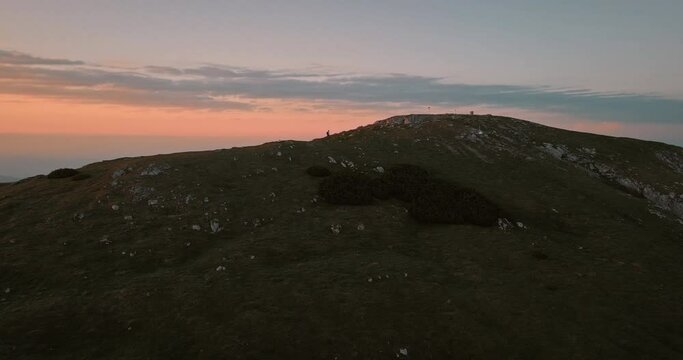Ad rone shot of the mountain Peca, in the distance a hiker is walking on the ridge. Orange sky colors with some clouds.