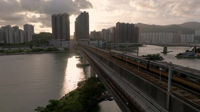 MTR train drives over a girder bridge towards Tsing Yi, Hong Kong. Backwards revealing drone shot during golden hour.