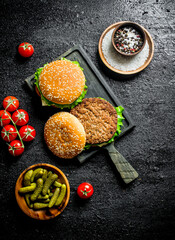 Burgers on a cutting Board with gherkins and spices in bowls.