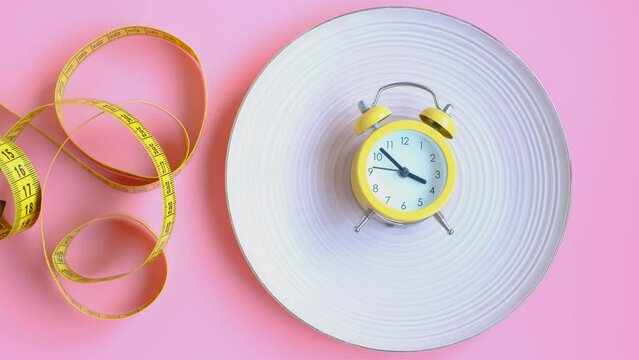 Plate with alarm clock and centimeter tape on pink background, intermittent fasting concept.