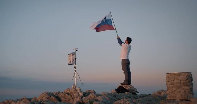 Man standing on a rock and holding a hiking pole in the air with attaches slovenian flag. Clear early morning sky with a touch of orange.