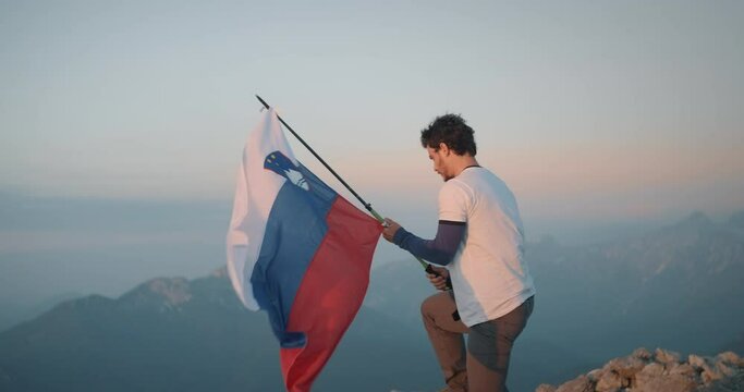 Man holding a hiking pole on which he attached a slovenian flag, steps on the rock and lets the flag flutter in the wind.