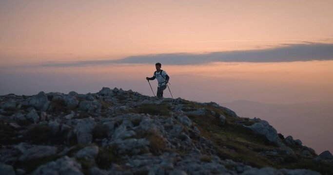 A camera tracking hiker frontal walkig up to the top of mountain Peca in the early morning. Orange color in the sky with some clouds.