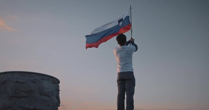 Camera circling aroud the hiker at the top of mountain Peca holding a pol with a slovenian flag attached on it, fluttering in the wind.