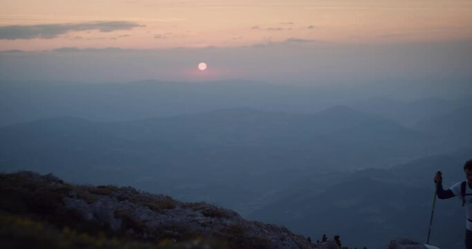Hiker with hiking poles and a backpack climbing up a mountain in the early morning. In backgroud sun is rising.