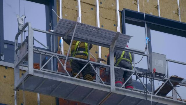 At a construction site, builders in a cradle on cables fastens decorative cladding panels on the wall of a building under construction