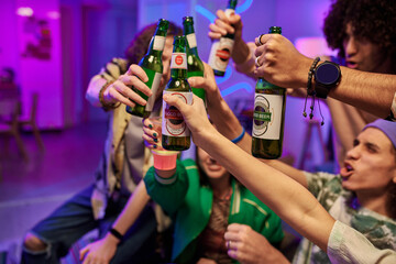 Close-up of hands of young intercultural people toasting with green bottles of beer at home party...