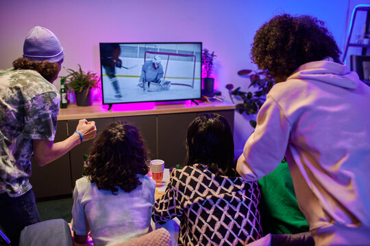 Rear View Of Friends Sitting In Front Of Tv Screen And Watching Broadcast Of Hockey Match In Living Room Lit By Neon Light At Leisure