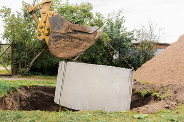 Excavator with a bucket, lowering into the pit on steel cables concrete sewer ring. Construction or...