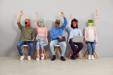 Group of people wearing funny Halloween animal masks sitting in row, holding digital devices,...