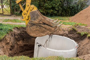 Excavator with a bucket, lowering into the pit on steel cables concrete sewer ring. Construction or...
