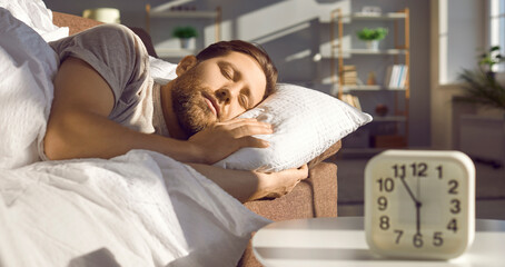 Man sleeping peacefully on the sofa in a sunny bedroom or living room with an alarm clock showing 6...