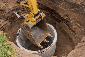 Excavator with a bucket, lowering into the pit on steel cables concrete sewer ring. Construction or repair of sewer home - obrazy, fototapety, plakaty