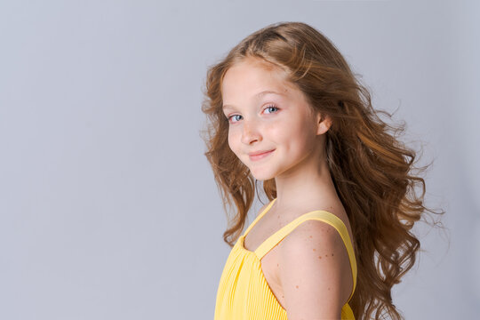 Happy half-length school age girl portrait in yellow clothes with long wavy blond hair smiles sweetly, isolated on light gray background. Caucasian girl model
