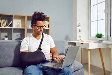 Young man with broken wrist wearing medical arm sling, sitting on sofa at home, working on laptop...